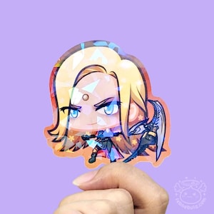 Op de afbeelding: Een holografische sticker met een stripfiguur met blond haar, blauwe ogen en een vastberaden uitdrukking. Het personage draagt een blauw en zwart harnas en houdt een wapen vast. De sticker heeft een kleurrijke, iriserende afwerking.