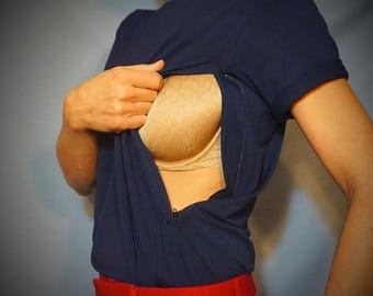 Puede incluir: Camiseta azul marino con una discreta abertura con cremallera, que revela un sujetador de encaje beige. La modelo lleva pantalones rojos. La imagen se centra en el diseño de la camiseta y el sujetador.