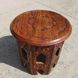 Puede incluir: Una mesa auxiliar redonda de madera con intrincadas tallas y un acabado marrón oscuro. La tapa presenta un diseño floral con detalles de latón. Los lados de la mesa tienen recortes geométricos abiertos. La mesa mide aproximadamente 50 cm de diámetro y 50 cm de alto.