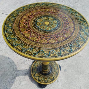 May include: A round, yellow side table with intricate, hand-painted floral and geometric patterns in black, gold, red, and teal. The table has a single pedestal base and measures approximately 24 inches in diameter.