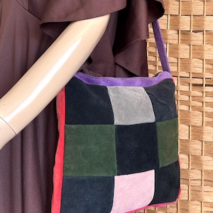 May include: A patchwork suede shoulder bag with a purple strap and trim. The bag features squares of black, grey, green, and pink suede. The bag is held by a mannequin wearing a brown dress.