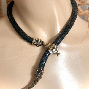 May include: Black snake-shaped necklace with gold-coloured metal accents. The necklace features a snake head and tail design with clear crystal accents. The snake's body is a textured black material.