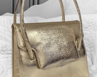 May include: A gold metallic handbag with a structured rectangular shape and two top handles. The bag has a subtle textured finish.