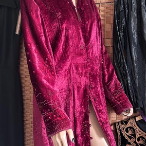 May include: A long, claret velvet coat with a mandarin collar and button fastenings. The coat features intricate beaded embroidery on the collar, cuffs, and front. The coat is displayed on a mannequin.