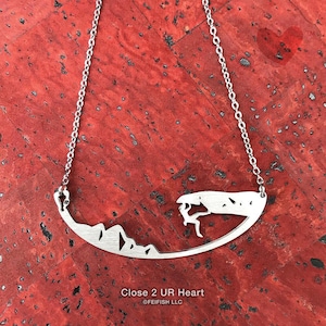 Peut inclure: Un collier en métal argenté avec un pendentif en forme de croissant présentant une découpe représentant une chaîne de montagnes et une personne faisant de l'escalade. Le pendentif est fixé à une fine chaîne. Le texte "Close 2 UR Heart" est imprimé en dessous.