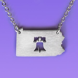 Puede incluir: Collar de plata con un recorte del estado de Pensilvania con la Campana de la Libertad en su interior. El collar está sobre un fondo morado. El texto "Close 2 UR Heart" y "©FEIFISH LLC" está en la parte inferior de la imagen.