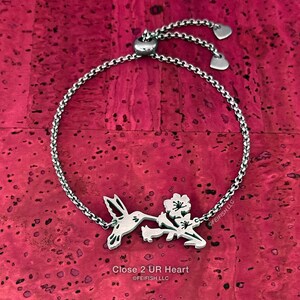 May include: A silver bracelet featuring a hummingbird and flower charm. The bracelet has a sliding clasp with two small hearts. The charm reads "Close 2 UR Heart" and "@FEIFISH LLC".