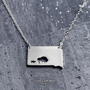 May include: Silver necklace with a cutout of the state of South Dakota, featuring a bison and a calf. The necklace has a chain and a pendant with the text "Close 2 UR Heart" and "©FEIFISH LLC".