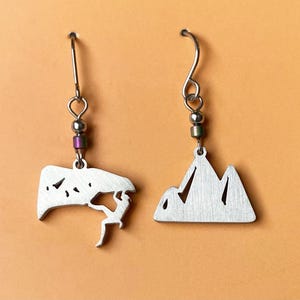 May include: A pair of silver earrings with a climber silhouette on one and a mountain silhouette on the other. The earrings have a silver bead and a purple or green bead on the hook.