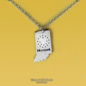 May include: Silver necklace with a pendant shaped like the state of Indiana. The pendant has a brushed finish and the word "Hoosier" is engraved on it. The necklace is on a yellow background.
