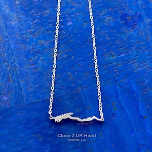 May include: A silver necklace with a mountain range pendant, suspended from a delicate chain. The necklace is displayed against a textured blue background. The text "Close 2 UR Heart" and the brand name "@FEIFISH LLC" are visible.