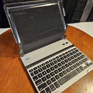 May include: A silver and black ZAGG keyboard case for a tablet. The keyboard has black keys with white lettering and symbols. The case is open, revealing a black interior and a silver frame. The ZAGG logo is visible on the keyboard.