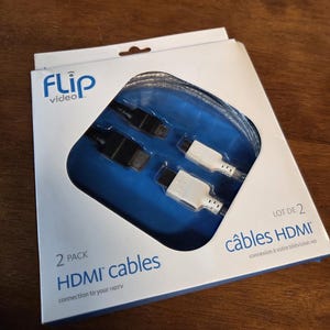 May include: A white and blue box containing two HDMI cables. The box has the "Flip video" logo and text that reads "2 PACK HDMI cables connection to your HDTV". The cables have black and white connectors.