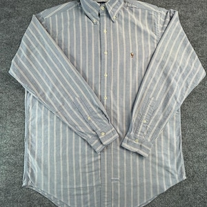 Op de afbeelding: Een lichtblauw overhemd met lange mouwen en verticale witte strepen, voorzien van een button-down kraag en een klein geborduurd logo op de linkerborst. Het overhemd heeft een knoopsluiting aan de voorzijde, knoopmanchetten en een afgeronde zoom.