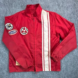 May include: A bright red zip-up windbreaker jacket featuring a vertical white stripe on the left side. The right sleeve displays two circular patches, one with a cartoon character and one reading National Hot Rod Association. A Dodge Racing Team patch is on the chest.
