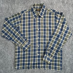 May include: A long-sleeve button-down shirt featuring a plaid pattern in shades of navy blue, olive green, and white. The shirt includes a pointed collar with button-down points, a single chest pocket, and white buttons running down the front placket.