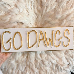 May include: A rectangular canvas with the words "GO DAWGS" in gold, raised lettering. The canvas is white with a gold border. The background is a soft, white, feathery texture.
