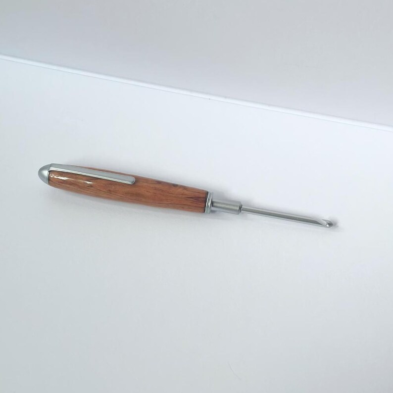 May include: A crochet hook with a silver metal hook and shaft. The handle is made of dark brown wood with a rounded silver metal tip. The hook is designed for yarn crafts, featuring a slender, ergonomic design for comfortable use. The hook is set against a white background.