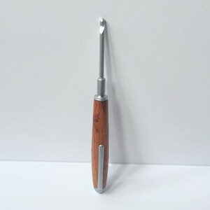 May include: A crochet hook with a silver metal hook and shaft. The handle is made of dark brown wood with a rounded silver metal tip. The hook is designed for yarn crafts, featuring a slender, ergonomic design for comfortable use. The hook is set against a white background.