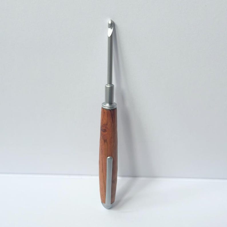 May include: A crochet hook with a silver metal hook and shaft. The handle is made of dark brown wood with a rounded silver metal tip. The hook is designed for yarn crafts, featuring a slender, ergonomic design for comfortable use. The hook is set against a white background.
