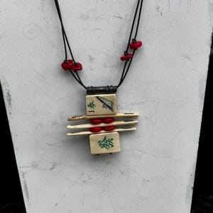 May include: A unique necklace featuring a pendant made of ivory-colored tiles with green and black symbols, accented by red beads. The necklace has a black cord and is displayed on a white stand.