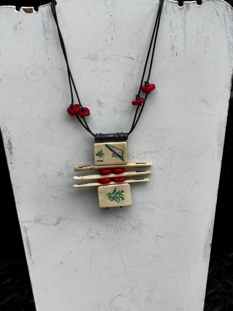 May include: A unique necklace featuring a pendant made of ivory-colored tiles with green and black symbols, accented by red beads. The necklace has a black cord and is displayed on a white stand.