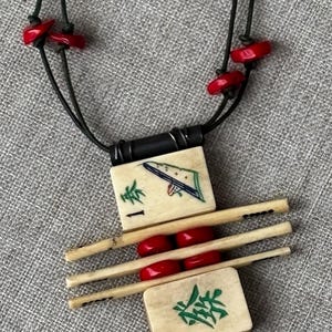 May include: A unique necklace featuring a pendant made of ivory-colored tiles and wooden sticks, accented with red beads. The tiles have green and blue painted designs and black cord. The necklace is on a gray textured surface.