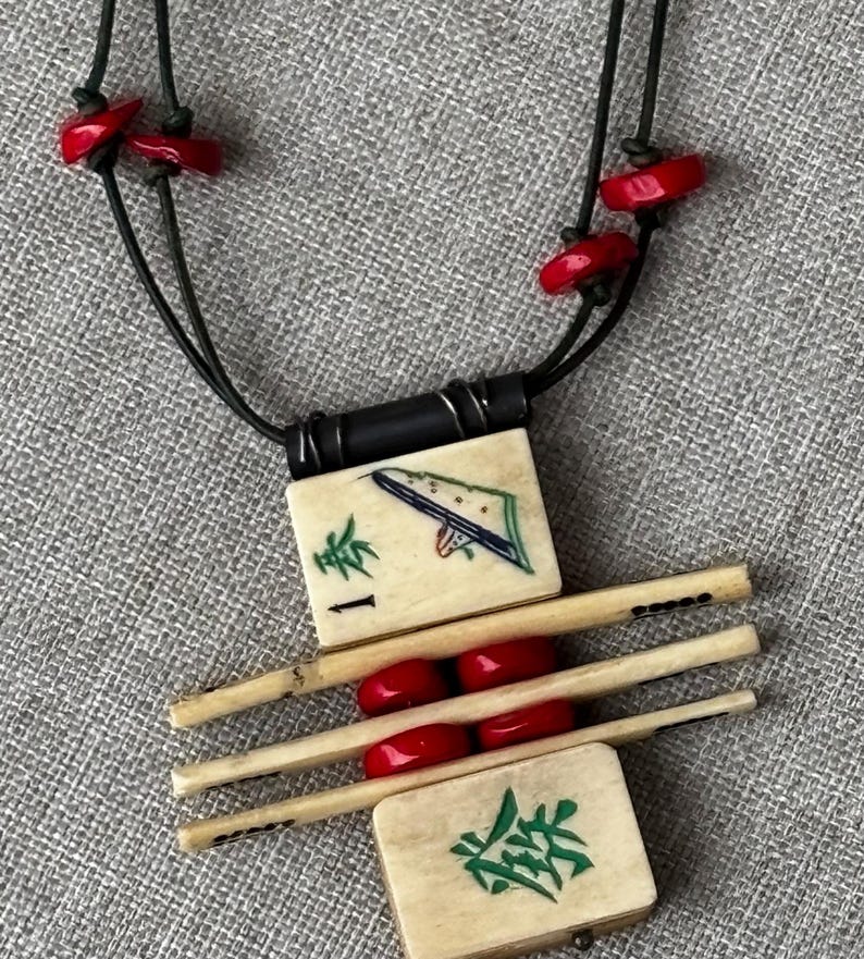 May include: A unique necklace featuring a pendant made of ivory-colored tiles and wooden sticks, accented with red beads. The tiles have green and blue painted designs and black cord. The necklace is on a gray textured surface.