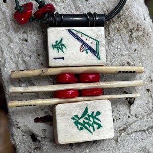 May include: A handcrafted necklace featuring a pendant made from vintage Mahjong tiles. The pendant has a black cord and is accented with red beads. The tiles display green and blue artwork and Chinese characters.