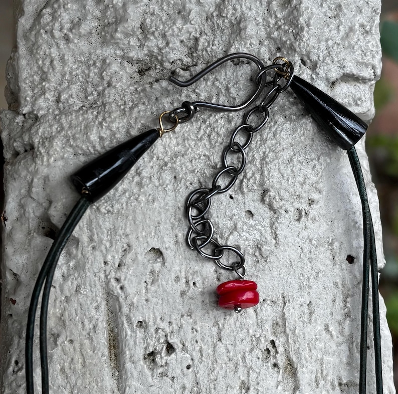 May include: A close-up of a necklace featuring black cone-shaped beads and a red bead pendant. The necklace has a silver-colored chain and a hook clasp. The necklace is displayed against a textured, light-colored background.