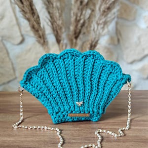 Peut inclure: Un sac à main en crochet en forme de coquille de couleur bleu turquoise avec une bandoulière en chaîne dorée et un petit charme en forme d'abeille.
