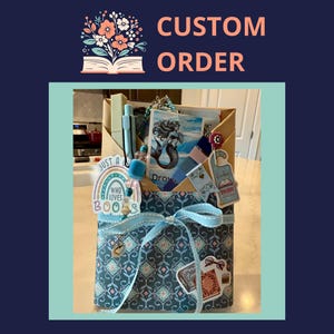 May include: A custom order gift box with a blue and white patterned exterior, tied with a light blue ribbon. The box contains a pen, mermaid art, and a "Just a Who Loves Books" sticker. The words "Custom Order" are at the top.