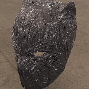 Puede incluir: Una máscara gris oscuro texturizada con un diseño detallado, con aberturas para los ojos y una forma contorneada. La máscara tiene una estética elegante y moderna, lo que sugiere un tema de superhéroe o cosplay.