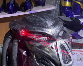 May include: A detailed, metallic Predator mask with glowing red eyes and intricate detailing. The mask is black and silver, with a textured surface. Other pieces of a costume are visible in the background, including blue and gold armor pieces.