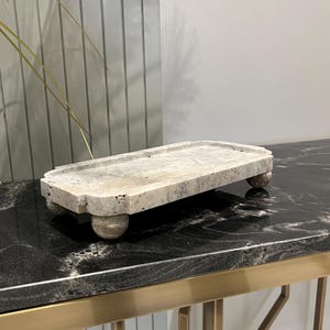 Könnte beinhalten: Ein rechteckiges Tablett aus hellem Graustein mit abgerundeten Kanten und vier kleinen Füßen. Das Tablett steht auf einer schwarzen Granitoberfläche.