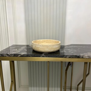 Puede incluir: Un cuenco de piedra beige descansa sobre una consola negra y dorada. La mesa tiene una tapa de m&aacute;rmol negro con ribetes dorados y una base geom&eacute;trica de metal dorado. El fondo es una pared blanca.