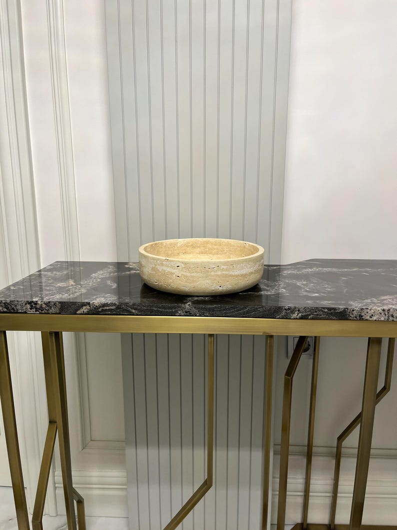 Puede incluir: Un cuenco de piedra beige descansa sobre una consola negra y dorada. La mesa tiene una tapa de m&aacute;rmol negro con ribetes dorados y una base geom&eacute;trica de metal dorado. El fondo es una pared blanca.