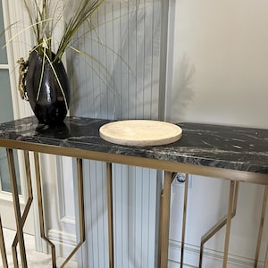 May include: A round, light beige marble serving tray sits on a black marble top console table with a gold metal frame. The table has a geometric design and is in a modern interior setting.