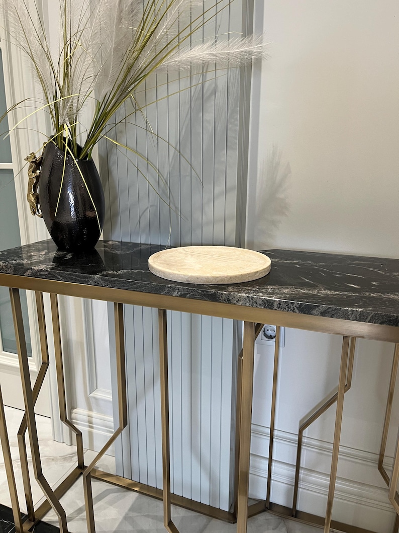 May include: A round, light beige marble serving tray sits on a black marble top console table with a gold metal frame. The table has a geometric design and is in a modern interior setting.