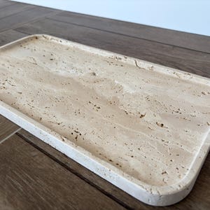 K&ouml;nnte beinhalten: Ein rechteckiges Tablett aus hellem beigefarbenem Travertinstein. Das Tablett hat einen leicht abgerundeten Rand und eine glatte, strukturierte Oberfl&auml;che.