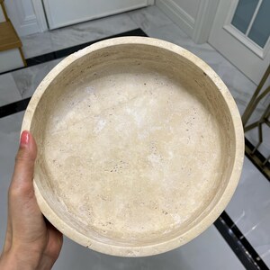 Puede incluir: Un cuenco redondo de piedra beige con interior liso y exterior texturizado. El cuenco est&aacute; sostenido por una mano, mostrando sus variaciones de color naturales y su forma circular. Este cuenco decorativo es adecuado para la decoraci&oacute;n del hogar.