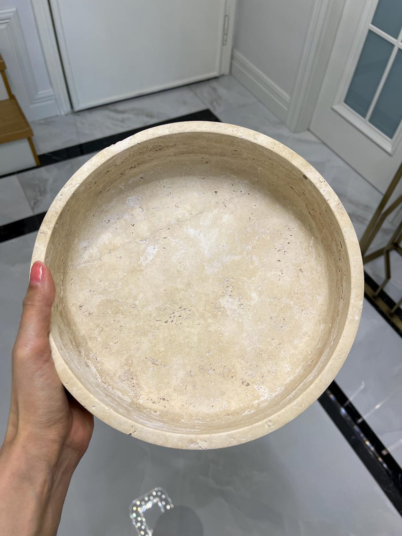 Puede incluir: Un cuenco redondo de piedra beige con interior liso y exterior texturizado. El cuenco est&aacute; sostenido por una mano, mostrando sus variaciones de color naturales y su forma circular. Este cuenco decorativo es adecuado para la decoraci&oacute;n del hogar.