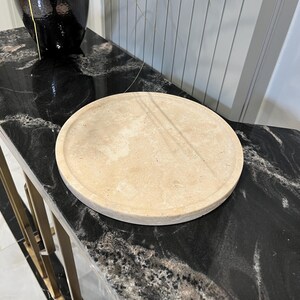 May include: A round, light beige, decorative tray made of natural stone. The tray is sitting on a black marble surface.
