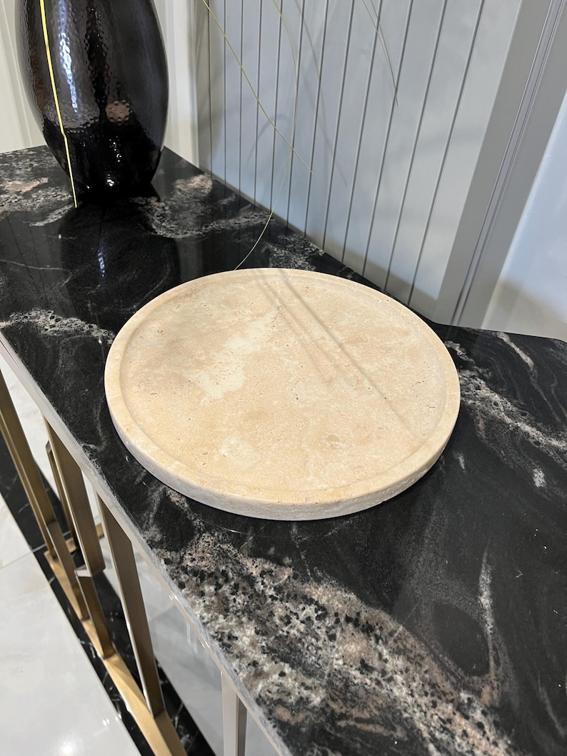May include: A round, light beige, decorative tray made of natural stone. The tray is sitting on a black marble surface.