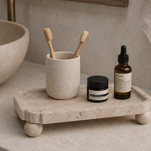 May include: A rectangular beige stone tray with four spherical feet, holding a matching stone toothbrush holder with two wooden toothbrushes, a small black jar of Aesop cream, and a brown glass dropper bottle with an Aesop label.
