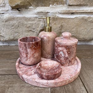Op de afbeelding: Een set badkameraccessoires van roze marmer. De set bevat een zeepdispenser met een gouden pomp, een beker, een pot met deksel, een klein schaaltje en een rond dienblad. Het marmer heeft een natuurlijk, geaderd patroon.