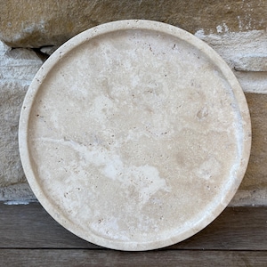 May include: A round, beige travertine coaster with a smooth, polished surface. The coaster has a natural, organic look with subtle variations in color and texture.