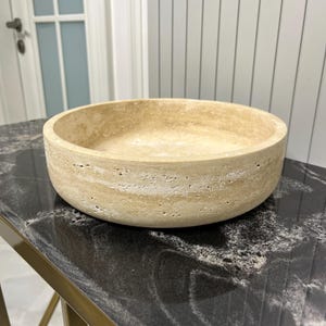 Puede incluir: Un cuenco de piedra beige con forma redondeada. El cuenco tiene una superficie texturizada y se apoya sobre una mesa negra y dorada. El cuenco es un objeto decorativo.