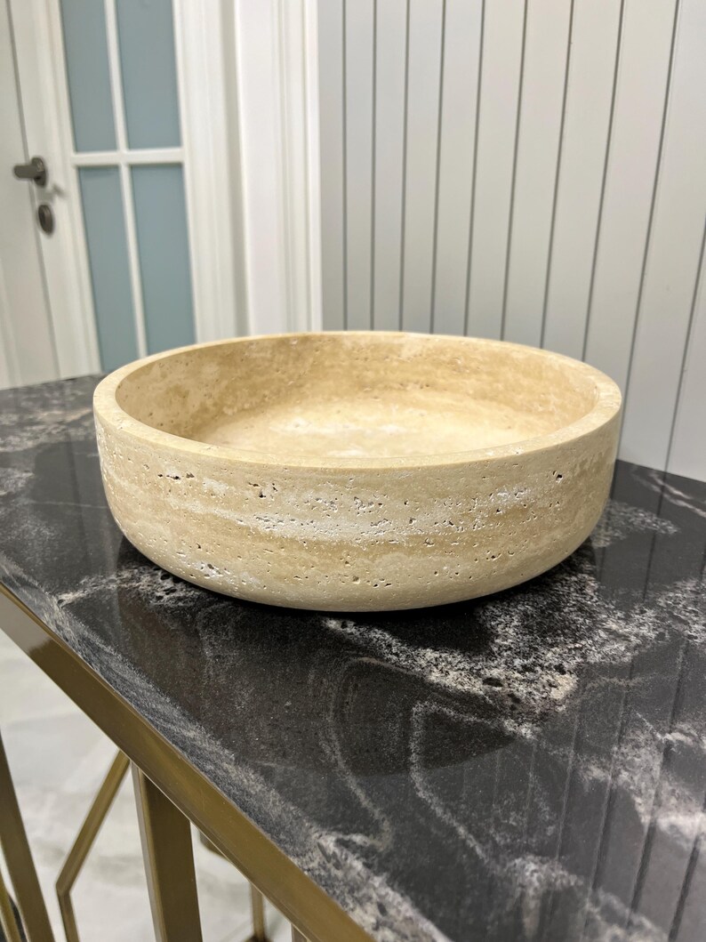 Puede incluir: Un cuenco de piedra beige con forma redondeada. El cuenco tiene una superficie texturizada y se apoya sobre una mesa negra y dorada. El cuenco es un objeto decorativo.