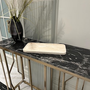 K&ouml;nnte beinhalten: Ein rechteckiges, hellbeiges Serviertablett aus Naturstein liegt auf einer schwarzen Granit-Arbeitsplatte mit goldenen Metallbeinen. Das Tablett befindet sich auf einem Konsolentisch mit geometrischem Design.